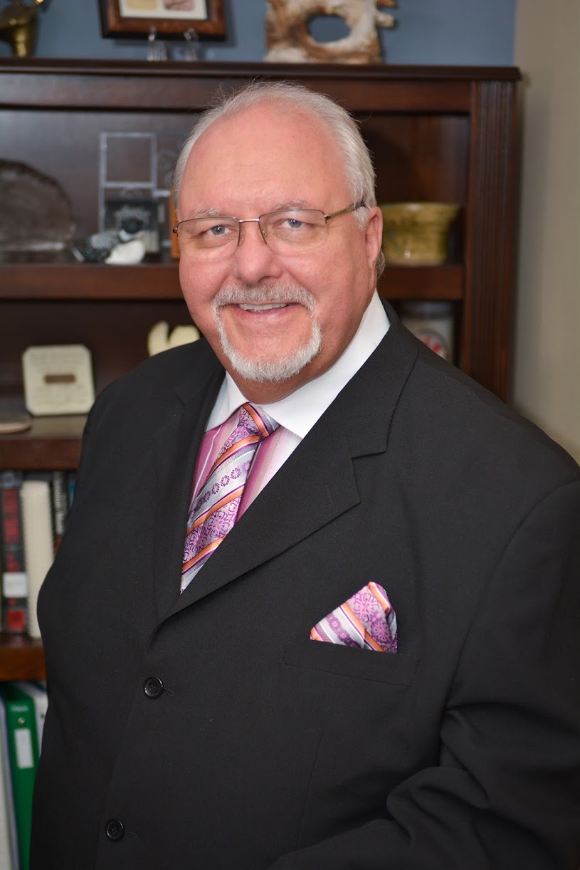 Dr. Jeffrey Frymire with a white shirt, black suit jacket matching purple orange and pink tie and handkerchief dark framed glasses.