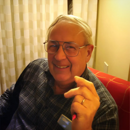 Ruben Schwieger man sitting in red chair smiling with dark framed glasses a wedding ring on and a blue plaid shirt.