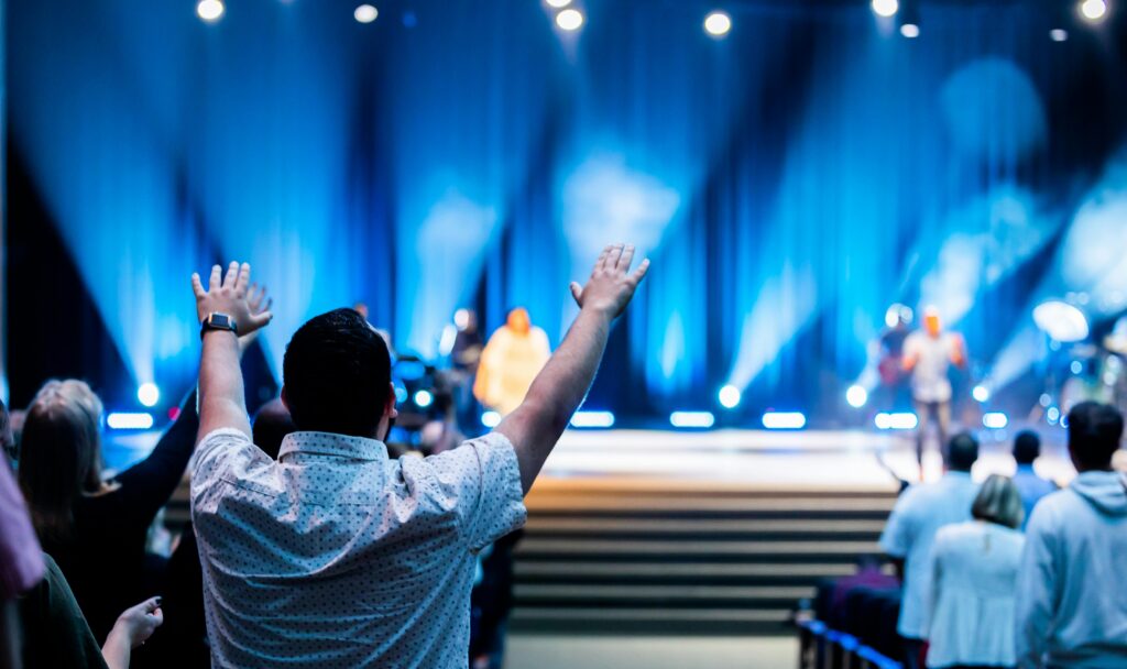 Hands raised in worship with lights on stage and a blurred band on stage with a man in the crowd in focus with hands up.