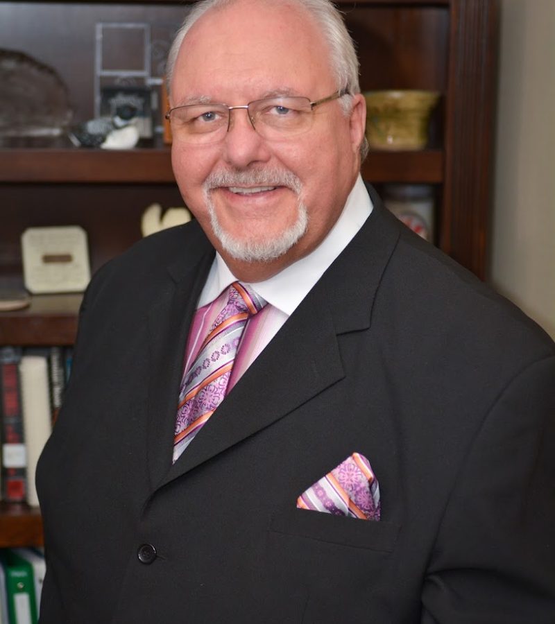 Dr. Jeffrey Frymire with a white shirt, black suit jacket matching purple orange and pink tie and handkerchief dark framed glasses.