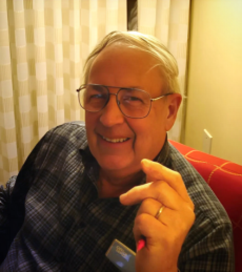 Ruben Schwieger man sitting in red chair smiling with dark framed glasses a wedding ring on and a blue plaid shirt.
