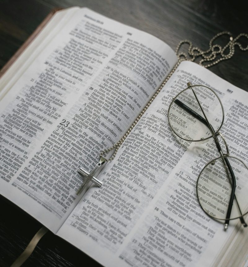 Bible open to Jeremiah 23 with a cross necklace laid down the fold and a pair of wired framed glasses laid folded on the right page.